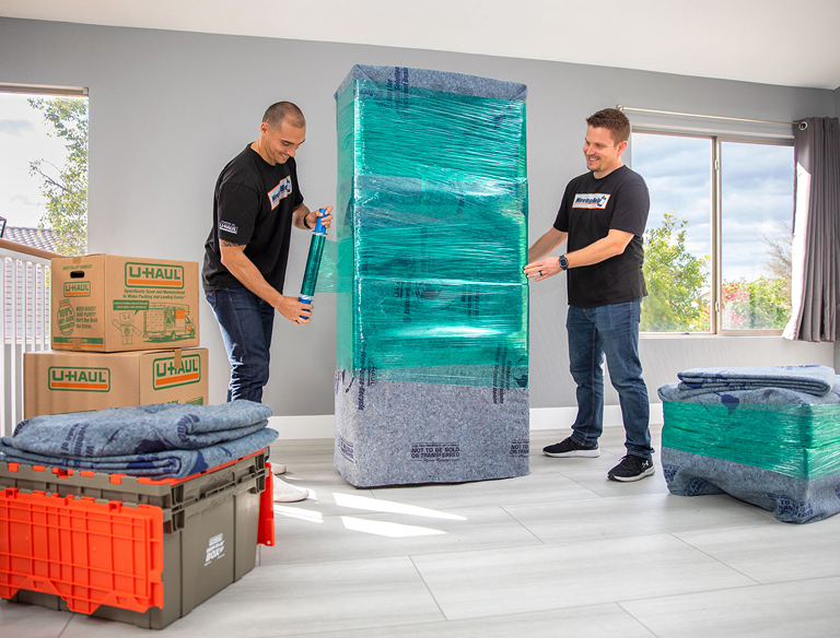Moving helpers wrapping furniture with stretch wrap next to U-Haul boxes and moving blankets