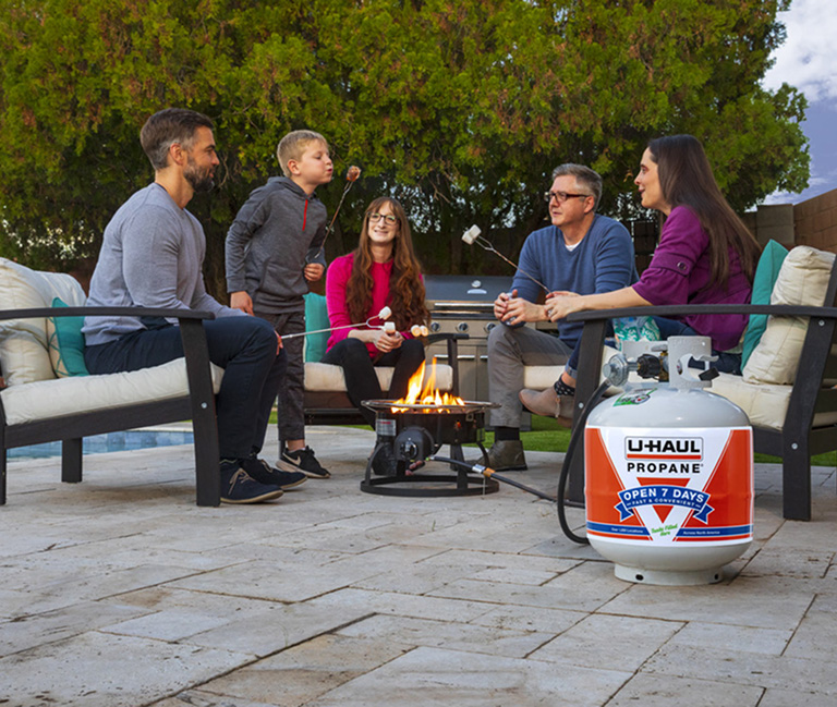 A family sits around a propane fire roasting marshmallows on skewers, watching their son blow the flame off his marshmallow