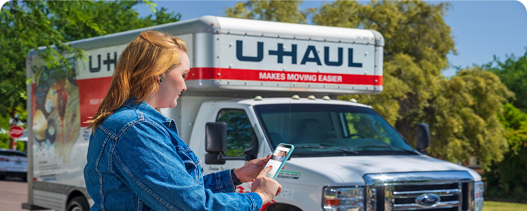 A woman standing near a U-Haul Moving Truck, using a mobile device to return her rental