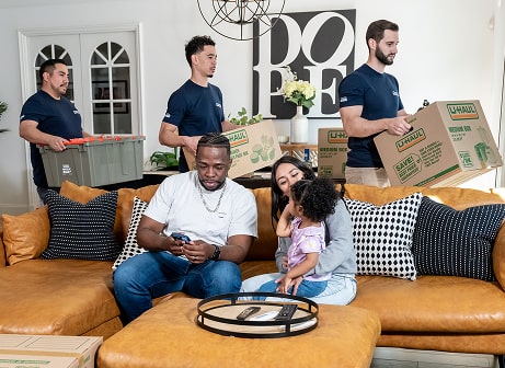 Moving helpers loading items into a family's home while the family sits on a couch