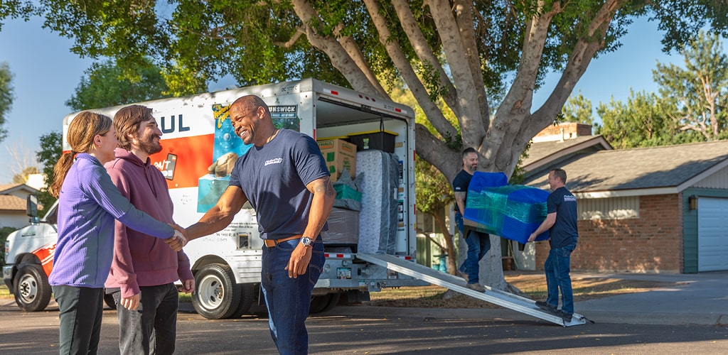 Moving Help® Service Providers loading heavy furniture into a U-Haul moving truck, while another service provider greets the family